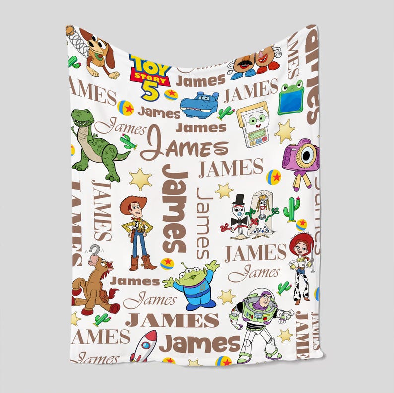 Puede incluir: Una manta de forro polar blanca con un patr&oacute;n repetitivo de personajes de Toy Story 5, incluyendo a Woody, Buzz Lightyear, Jessie, Rex y Slinky Dog, intercalados con el nombre James en varias fuentes marrones, estrellas amarillas y cactus verdes.