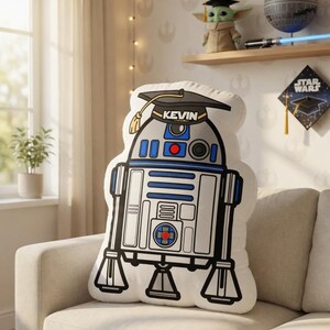 May include: A white R2-D2 shaped pillow with blue and red accents, wearing a graduation cap with the name "KEVIN". The pillow is on a beige couch. The room has Star Wars themed decor.