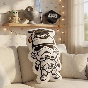 May include: A white and black Stormtrooper pillow wearing a graduation cap with the name "MARK" printed on it. The pillow is on a beige couch. A shelf in the background displays Star Wars themed decor.