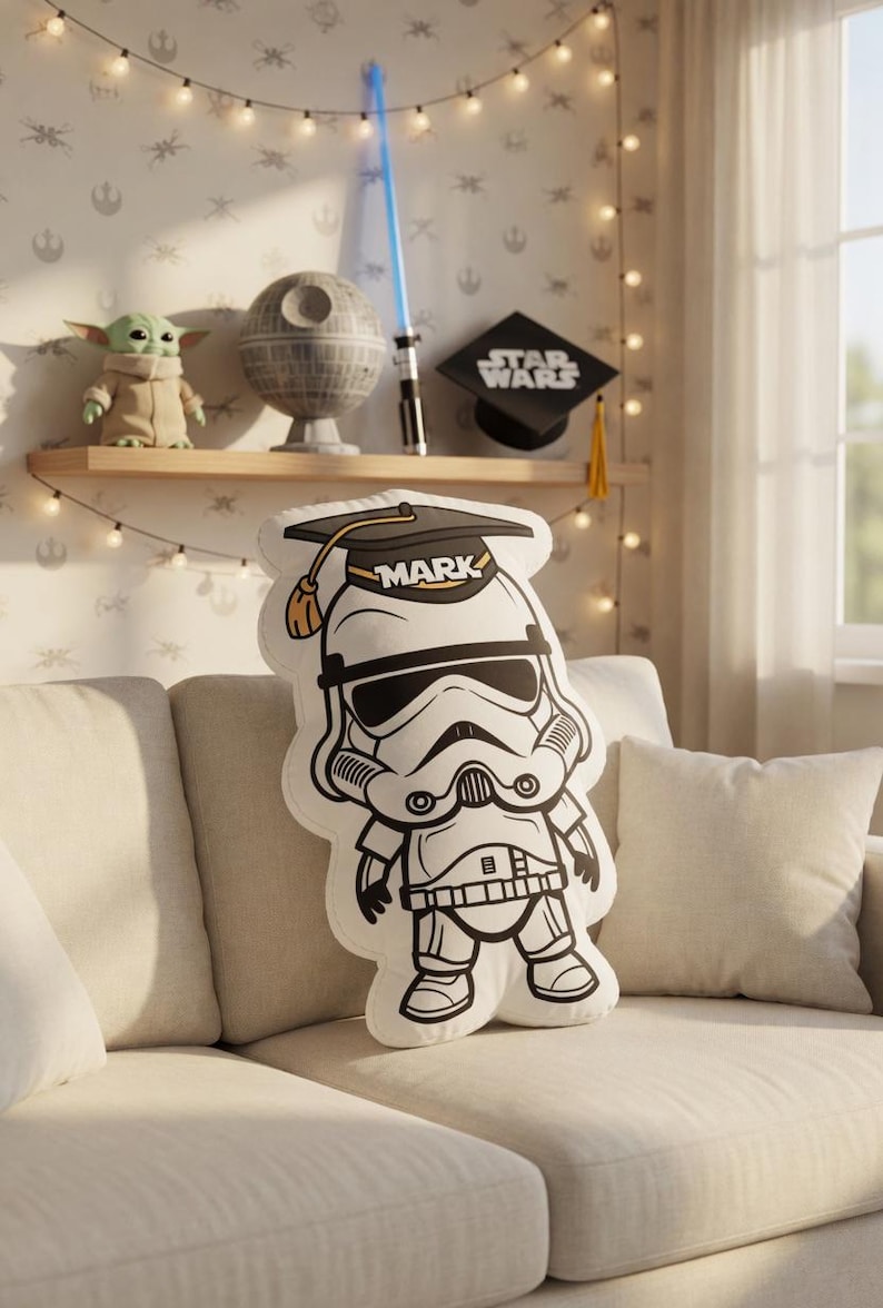 May include: A white and black Stormtrooper pillow wearing a graduation cap with the name "MARK" printed on it. The pillow is on a beige couch. A shelf in the background displays Star Wars themed decor.