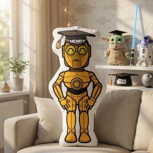 May include: A C-3PO shaped pillow wearing a graduation cap with the name "HENRY" on it. The pillow is yellow and black, and is on a beige couch. Other Star Wars themed items are on a shelf in the background.