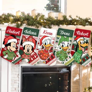 May include: Five Christmas stockings featuring Disney characters such as Mickey and Minnie Mouse, Donald and Daisy Duck, and Pluto. Each stocking has a name at the top and is decorated in red, green, and white festive patterns. The stockings are hung above a fireplace.