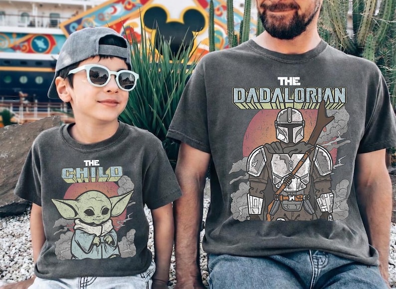 May include: Two charcoal gray t-shirts featuring graphic prints. The smaller shirt displays a green creature with the text THE CHILD. The larger shirt features a armored character with a staff and the text THE DADALORIAN. Both shirts have a vintage-inspired design with a circular background element.