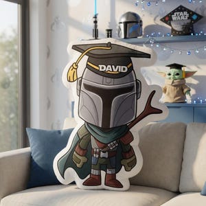 May include: A large, cartoon-style pillow shaped like a Mandalorian character wearing a graduation cap with the name "DAVID". The pillow is gray, green, and brown, with a yellow tassel. A Baby Yoda figure and Star Wars graduation caps are visible in the background.