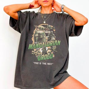May include: Dark gray t-shirt featuring a graphic from the Star Wars series, "The Mandalorian and Grogu." The design includes images of the Mandalorian, Grogu, and other characters, with the phrase "THIS IS THE WAY" at the bottom.