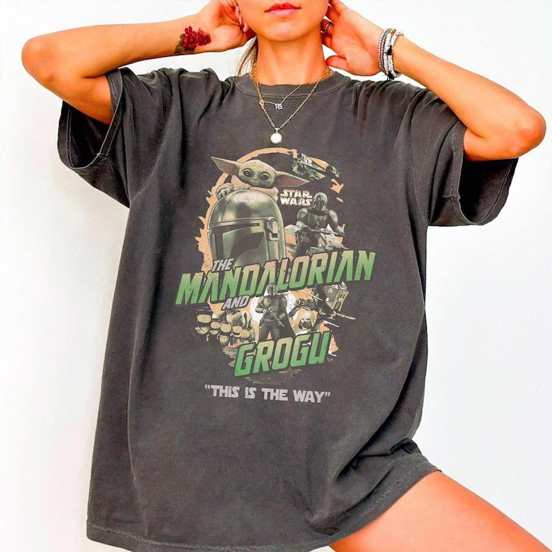 May include: Dark gray t-shirt featuring a graphic from the Star Wars series, "The Mandalorian and Grogu." The design includes images of the Mandalorian, Grogu, and other characters, with the phrase "THIS IS THE WAY" at the bottom.