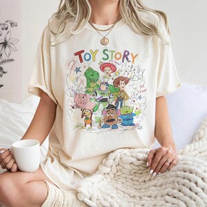 May include: Cream-colored t-shirt featuring a colorful graphic of Toy Story characters, including Woody, Buzz Lightyear, and Rex. The words "Toy Story" are written above the characters in a playful, multi-colored font. The design has a hand-drawn aesthetic.