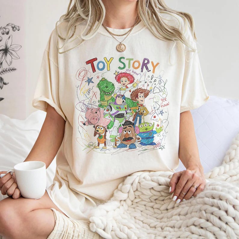 May include: Cream-colored t-shirt featuring a colorful graphic of Toy Story characters, including Woody, Buzz Lightyear, and Rex. The words "Toy Story" are written above the characters in a playful, multi-colored font. The design has a hand-drawn aesthetic.