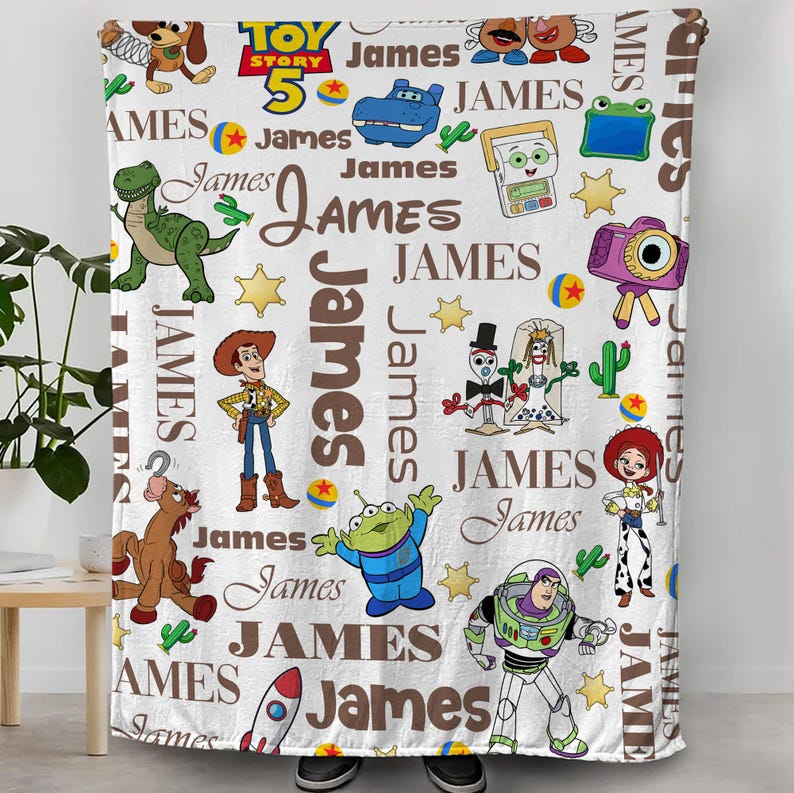 Puede incluir: Una manta de forro polar blanca con un patr&oacute;n repetitivo de personajes de Toy Story, incluyendo a Woody, Buzz Lightyear, Rex y el Alien, intercalados con el nombre James en letras marrones, estrellas amarillas y bolas de colores.