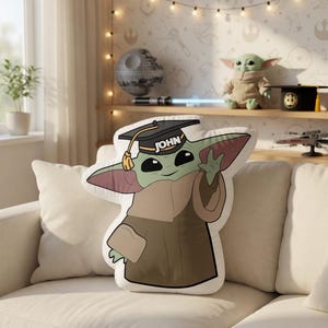May include: A plush pillow shaped like Grogu, also known as Baby Yoda, wearing a graduation cap with the name "JOHN" on it. The pillow is green, brown, and tan, and is sitting on a white couch. The background includes a shelf with Star Wars themed items.