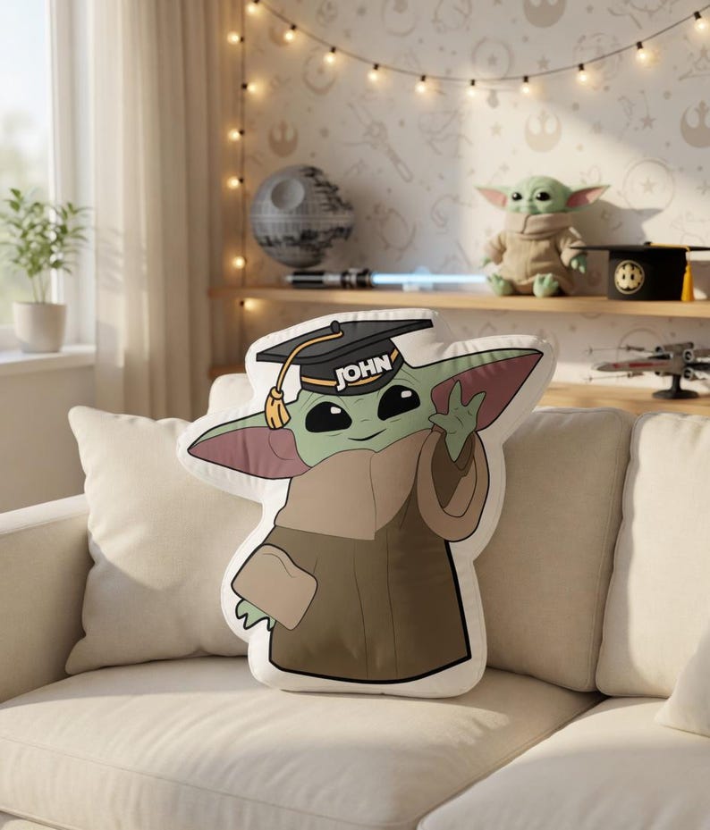 May include: A plush pillow shaped like Grogu, also known as Baby Yoda, wearing a graduation cap with the name "JOHN" on it. The pillow is green, brown, and tan, and is sitting on a white couch. The background includes a shelf with Star Wars themed items.