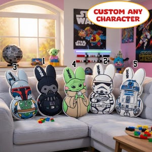 May include: Five Star Wars character-themed bunny cushions are displayed on a gray sofa. The cushions feature characters like Darth Vader, a Stormtrooper, and Baby Yoda. The cushions have the word "NAME" on the ears. The image also includes a Death Star model and Star Wars posters.