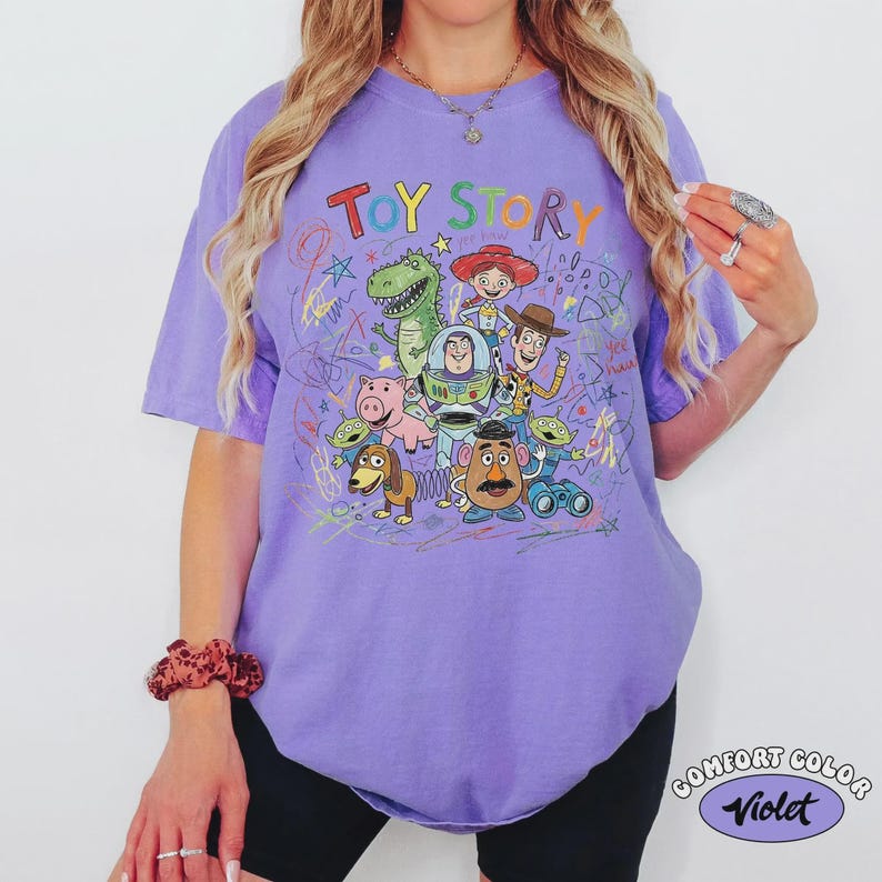 May include: A violet Comfort Colors t-shirt featuring a colorful graphic of Toy Story characters. The design includes the words "TOY STORY" above the characters, with a variety of cartoon figures. The shirt has short sleeves.
