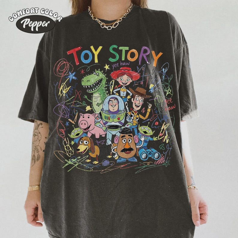 May include: Dark gray Comfort Colors t-shirt featuring a colorful graphic of Toy Story characters. The design includes the words "TOY STORY" in bright colors, along with images of Woody, Buzz Lightyear, and other characters.