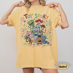 May include: Mustard yellow t-shirt featuring the words "TOY STORY" in colorful letters, along with cartoon characters like Woody, Buzz Lightyear, and Rex. The shirt has a Comfort Color label.