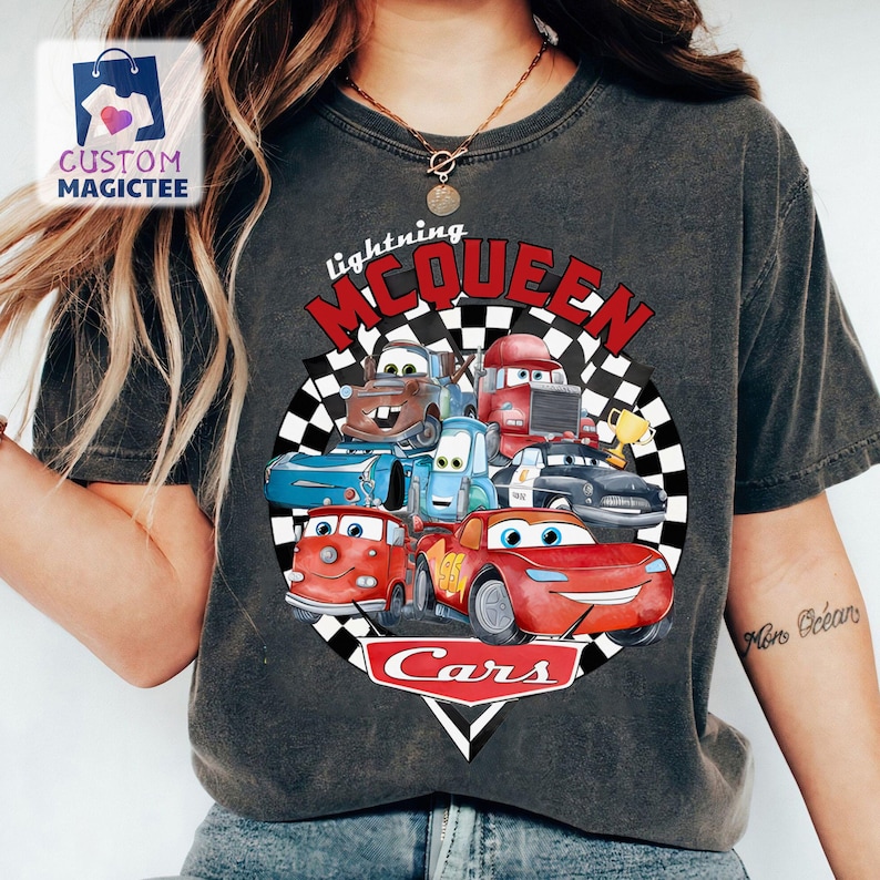 May include: Dark grey t-shirt with a colourful graphic of characters from the animated film Cars. The design includes Lightning McQueen and other vehicles, with the text "Lightning McQueen" and "Cars".