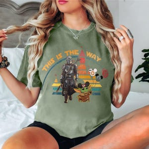 May include: Green t-shirt with a vintage-style graphic of a Mandalorian character and a Grogu character. The graphic includes the text "This is the way" and Mickey Mouse ears.