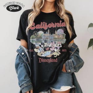 May include: Black t-shirt with a vintage Disneyland graphic. The design features the word "California" in pink, a castle, fireworks, and Disney characters. The text "Est. 1955" and "Disneyland" are also included.