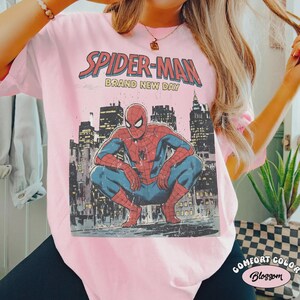 May include: Pink t-shirt featuring a vintage Spider-Man graphic. The design includes the words "Spider-Man Brand New Day" above an illustration of Spider-Man crouching in front of a city skyline. The shirt is a Comfort Colors brand.
