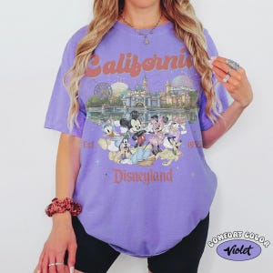 May include: A violet Comfort Color t-shirt featuring a Disneyland graphic. The graphic includes the text "California Disneyland Est. 1955" with images of Disney characters and park attractions. The shirt has short sleeves.
