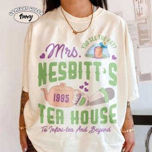 May include: Ivory Comfort Colors t-shirt with a graphic design. The design includes the text "Mrs. Nesbitt's Tea House 1995" in green and purple, along with a teapot and other illustrations. The phrase "To Infini-tea And Beyond" is also included.