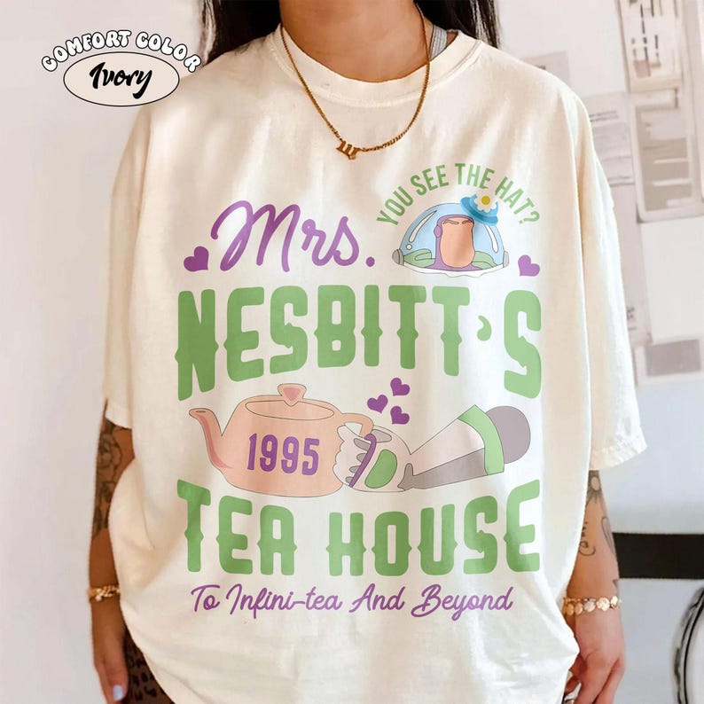 May include: Ivory Comfort Colors t-shirt with a graphic design. The design includes the text "Mrs. Nesbitt's Tea House 1995" in green and purple, along with a teapot and other illustrations. The phrase "To Infini-tea And Beyond" is also included.