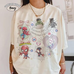 May include: Ivory-colored t-shirt featuring cartoon illustrations of Spidey and his Amazing Friends characters. The design includes Spider-Man, Hulk, Black Panther, and Spider-Gwen, with the words "Spidey and Amazing Friends" printed on the shirt.
