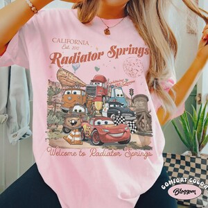May include: Pink t-shirt with a graphic of characters from the movie Cars, including Lightning McQueen. The shirt has the text "CALIFORNIA Est. 2012 Radiator Springs" and "Welcome to Radiator Springs". The shirt is a Comfort Color.