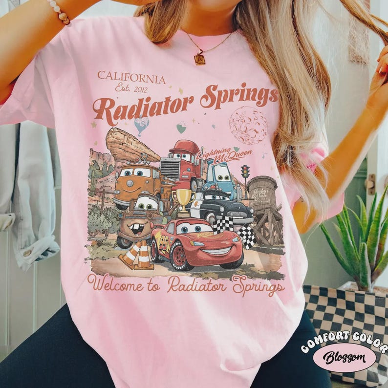 May include: Pink t-shirt with a graphic of characters from the movie Cars, including Lightning McQueen. The shirt has the text "CALIFORNIA Est. 2012 Radiator Springs" and "Welcome to Radiator Springs". The shirt is a Comfort Color.
