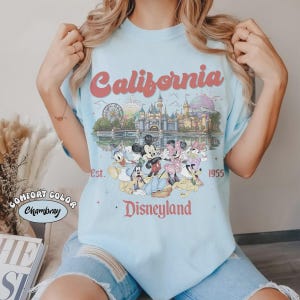 May include: Light blue t-shirt featuring a Disneyland design. The graphic includes the word "California" in a retro font, a castle, and various Disney characters. The text "Est. 1955" and "Disneyland" are also present.