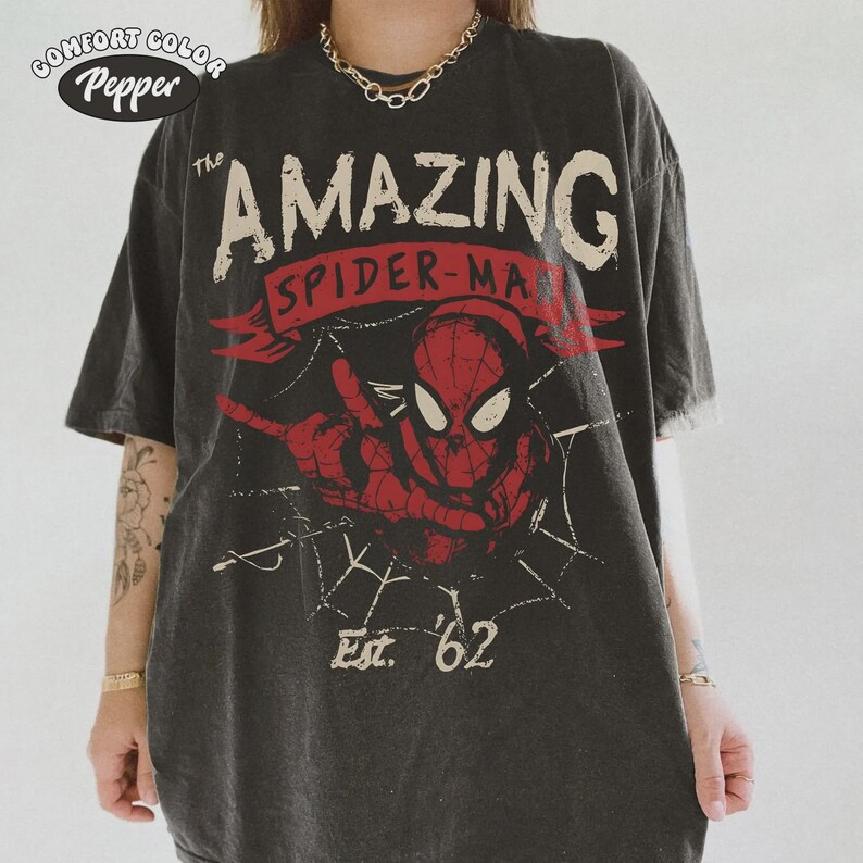 May include: Dark gray t-shirt featuring a distressed graphic of Spider-Man with the text "The Amazing Spider-Man" and "Est. '62". The design includes a red Spider-Man image and a web pattern. The shirt has a vintage look.