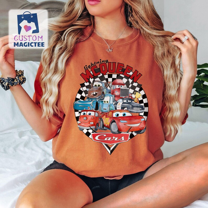 May include: Burnt orange t-shirt with a graphic of Lightning McQueen and other Cars characters. The design includes a checkered flag background and the word "Cars" in a white triangle. The shirt has a classic crew neck.