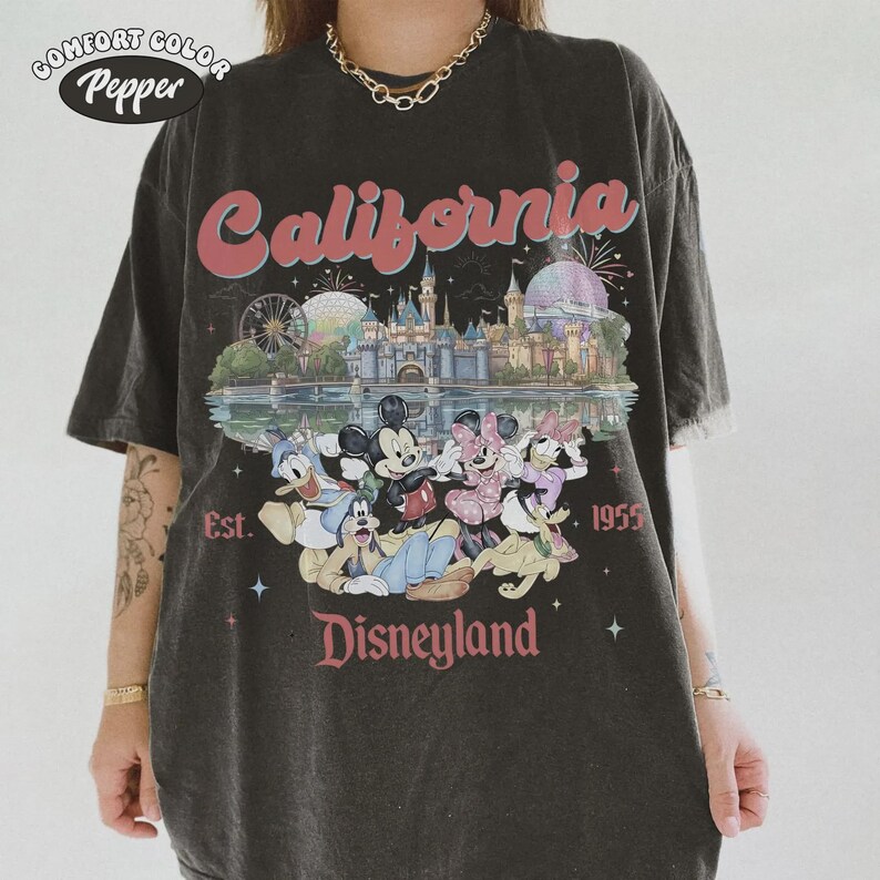May include: Dark gray t-shirt with a Disneyland graphic. The graphic features the word "California" in pink script above an illustration of Disneyland with Mickey Mouse, Minnie Mouse, Donald Duck, and Goofy. The text "Est. 1955" and "Disneyland" are also included.