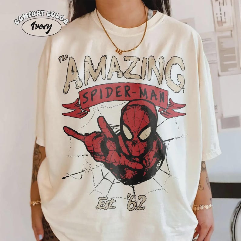 May include: Ivory Comfort Color t-shirt featuring a vintage-style graphic of Spider-Man. The design includes the text "The Amazing Spider-Man" above a red and black illustration of the superhero, with "Est. '62" below. The shirt has a classic, relaxed fit.