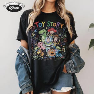 May include: Black Comfort Colors t-shirt featuring a colorful graphic of Toy Story characters. The design includes the words "TOY STORY" in rainbow colors, with "yee haw" accents. The shirt has a crew neck and short sleeves.