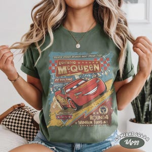 May include: Moss green t-shirt featuring a graphic of Lightning McQueen from the movie Cars. The design includes the text "Lightning McQueen" and "Round 4 Winner Takes All" in a vintage style. The shirt is labeled "Comfort Color".