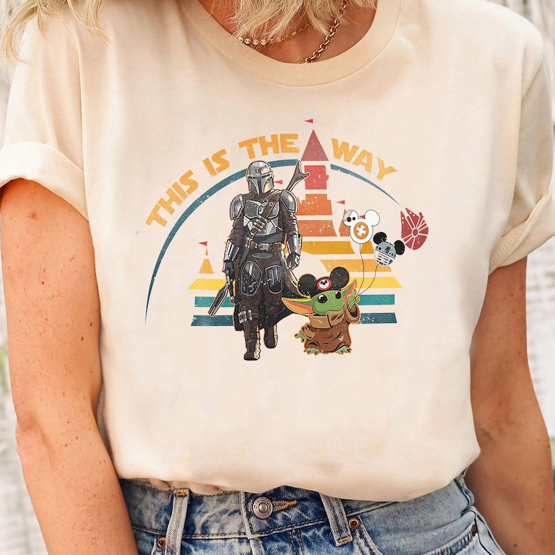 May include: A white t-shirt with a retro-style graphic of a Mandalorian character and a Grogu character in front of a castle. The text "This is the way" is printed in a curved arc above the characters.