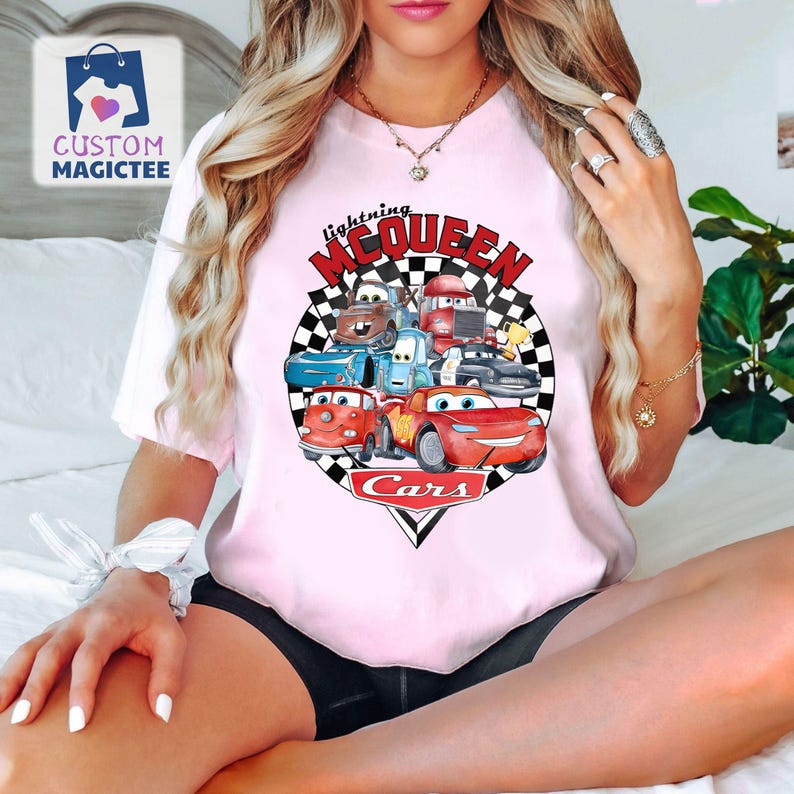 May include: Pale pink t-shirt with a graphic of Cars movie characters, including Lightning McQueen. The design is set within a black and white chequered circle, with the words "Lightning McQueen" and "Cars" in red.