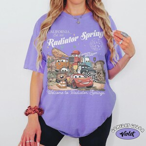 May include: A violet Comfort Colors t-shirt featuring a graphic of characters from the movie Cars, with the text "California Est. 2012 Radiator Springs" and "Welcome to Radiator Springs".