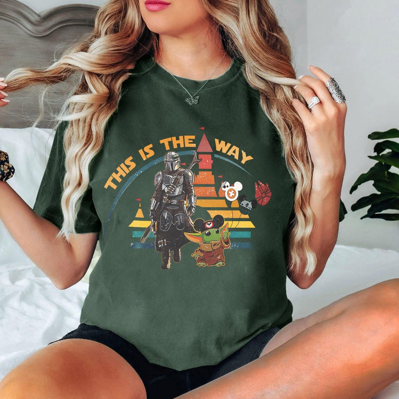 May include: A dark green t-shirt with a graphic print of a Star Wars character, a Mandalorian, and a Baby Yoda character. The text "This is the way" is printed on the shirt.