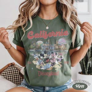 May include: Moss green t-shirt featuring a Disneyland California design. The design includes the word "California" in a pink font, a castle, and various Disney characters. The shirt is labeled "Comfort Color Moss".