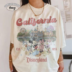 May include: Ivory-colored t-shirt with a Disneyland graphic. The design features the word "California" in pink, a castle, and cartoon characters. The text "Est. 1955" and "Disneyland" are also visible.
