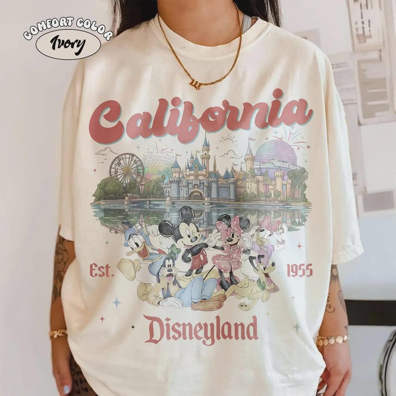 May include: Ivory-colored t-shirt with a Disneyland graphic. The design features the word "California" in pink, a castle, and cartoon characters. The text "Est. 1955" and "Disneyland" are also visible.
