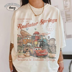 May include: Ivory-colored t-shirt featuring a graphic of characters from the movie Cars, with the text "CALIFORNIA Est. 2012 Radiator Springs" and "Welcome to Radiator Springs". The shirt has a vintage, distressed design.
