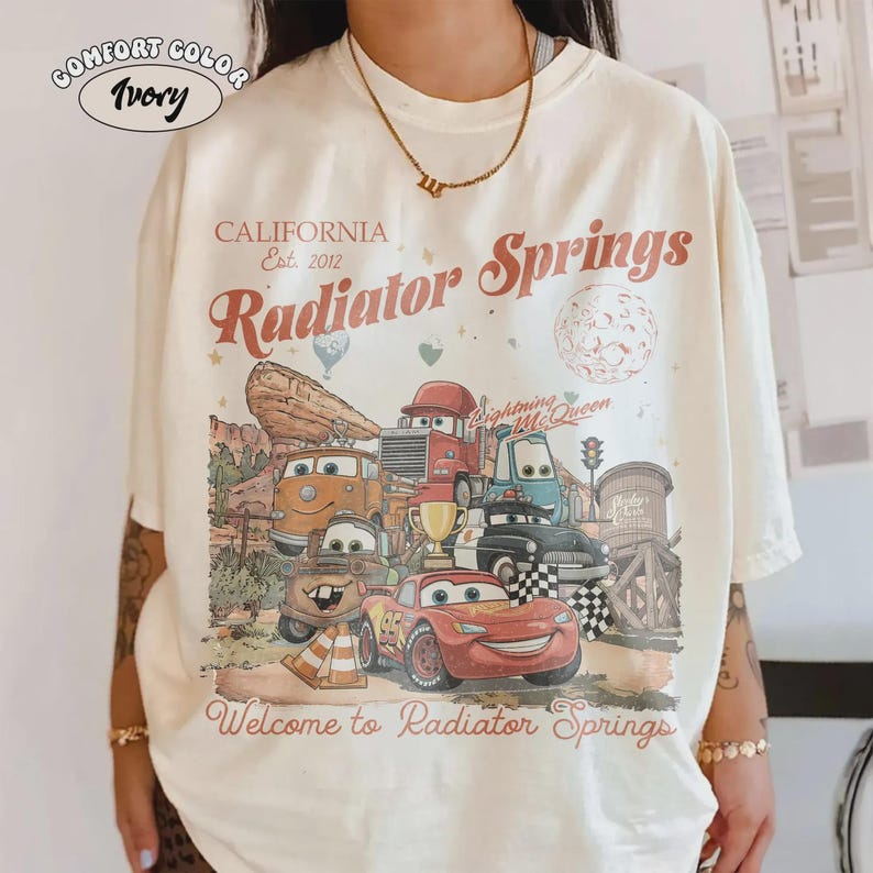 May include: Ivory-colored t-shirt featuring a graphic of characters from the movie Cars, with the text "CALIFORNIA Est. 2012 Radiator Springs" and "Welcome to Radiator Springs". The shirt has a vintage, distressed design.