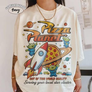 May include: Ivory-colored t-shirt with a Pizza Planet graphic. The design features a rocket, pizza, aliens, and the text "Pizza Planet." Additional text reads "Out of this world quality" and "Serving your local star cluster."