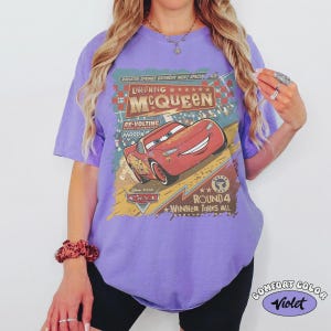 May include: A violet-colored t-shirt featuring a graphic of Lightning McQueen from the Disney Cars movie. The design includes text that reads "Lightning McQueen" and "Round 4 Winner Takes All."