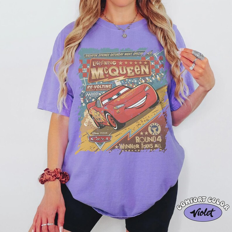 May include: A violet-colored t-shirt featuring a graphic of Lightning McQueen from the Disney Cars movie. The design includes text that reads "Lightning McQueen" and "Round 4 Winner Takes All."