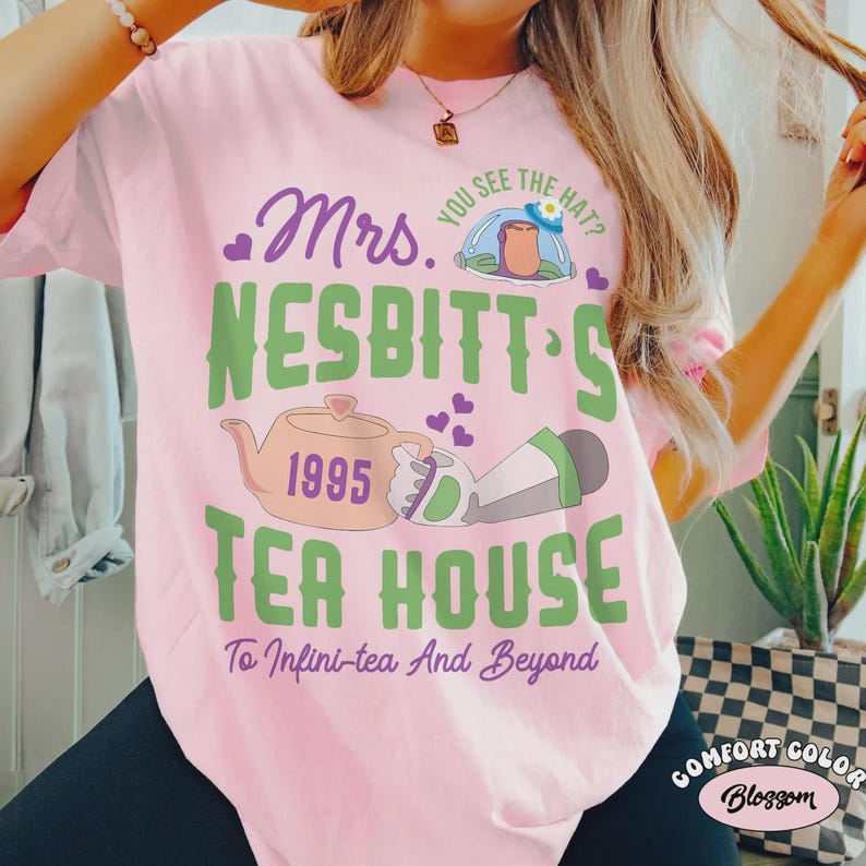 May include: A light pink t-shirt with a graphic design. The design includes the text "Mrs. Nesbitt's Tea House" with a teapot, the year "1995", and the phrase "To Infini-tea And Beyond". The shirt is a Comfort Colors Blossom.