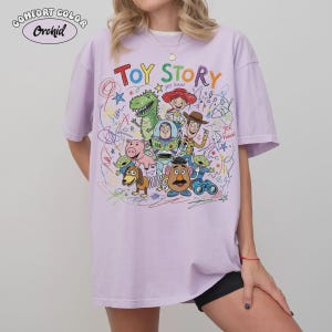 May include: A light purple Comfort Colors t-shirt featuring the words "Toy Story" in colorful letters. The shirt showcases a vibrant graphic of characters from the animated film, including Woody, Buzz Lightyear, and Rex.